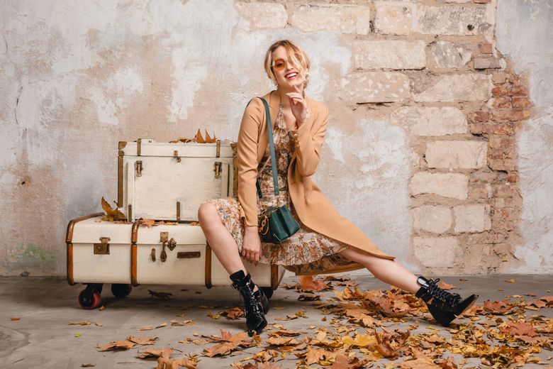 attractive stylish blonde woman in beige coat sitting on suitcases against wall in street, autumn fashion trend, wearing dress, boots, handbag, sunglasses, leaves on the ground, urban style Cizmele de cowboy, vedetele toamnei 2026. 5 ținute în care să le integrezi